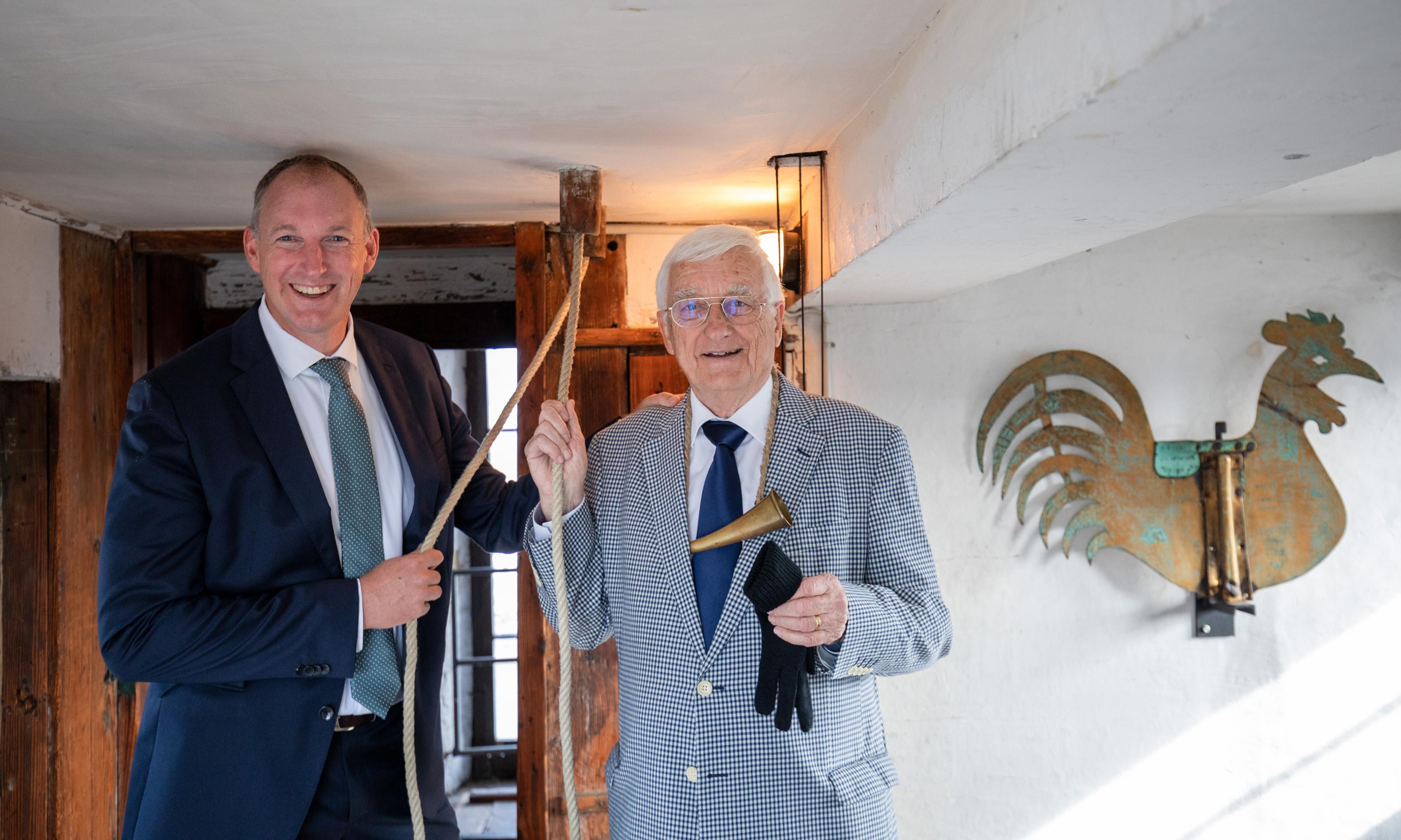 Foto des Messeglöckners Franz Bauer, in schwarzem Anzug mit gelber Kravatte gekleidet, mit dem Horn (Instrument) um den Hals gehängt und dem Seil zur Martinsglocke in den Händen.