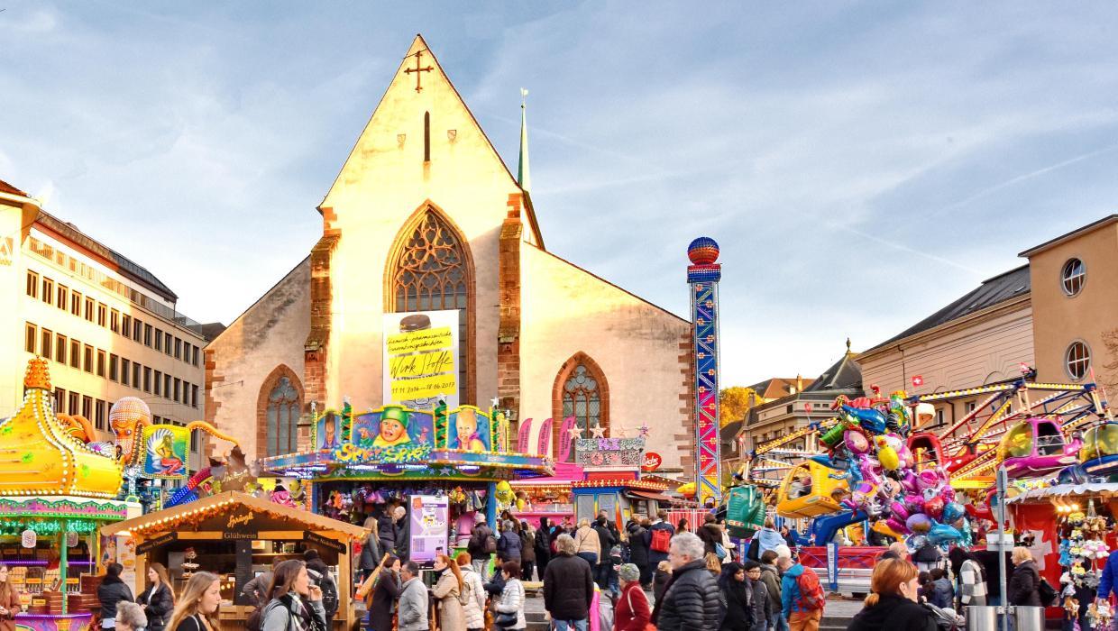 View of the vibrant hustle and bustle on Basel's Barfüsserplatz, with colourful shooting galleries, rides, food stands and the Barfüsserkirche in the background.