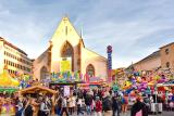 View of the vibrant hustle and bustle on Basel's Barfüsserplatz, with colourful shooting galleries, rides, food stands and the Barfüsserkirche in the background.