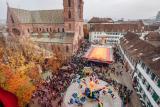 View from above the Münsterplatz with its autumn-coloured horse chestnut grove and the red Basel Minster. Colourful Autumn Fair rides spin in the square.