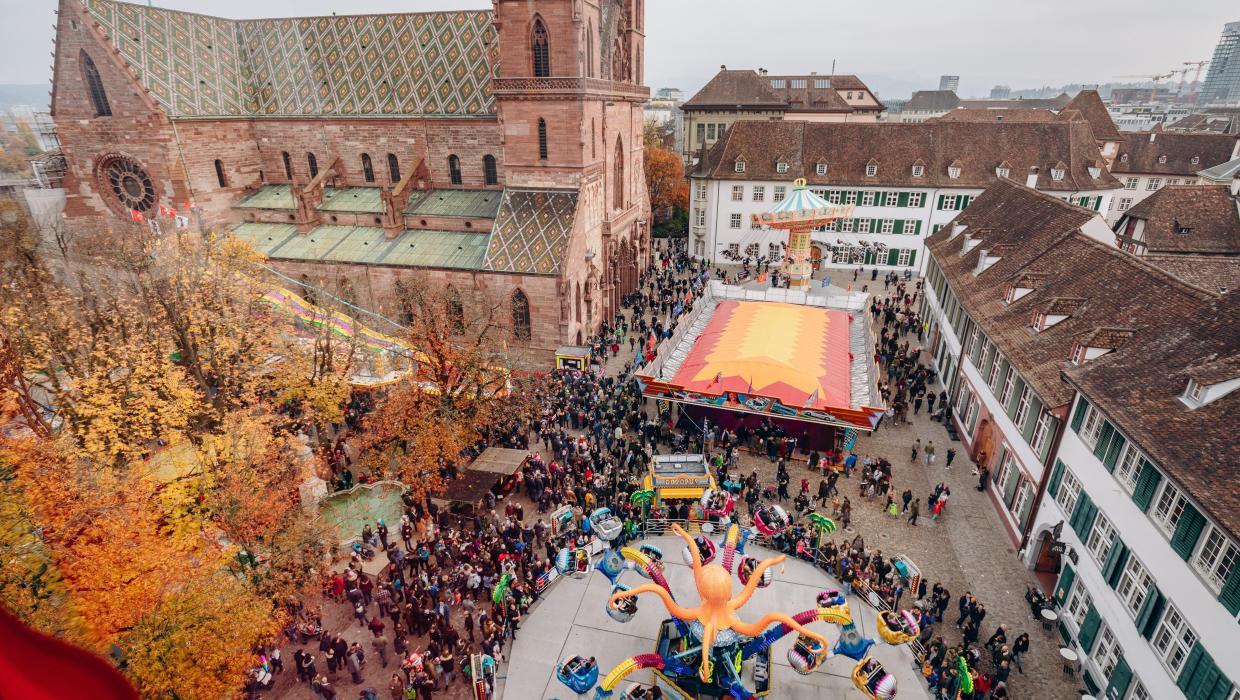 View from above the Münsterplatz with its autumn-coloured horse chestnut grove and the red Basel Minster. Colourful Autumn Fair rides spin in the square.